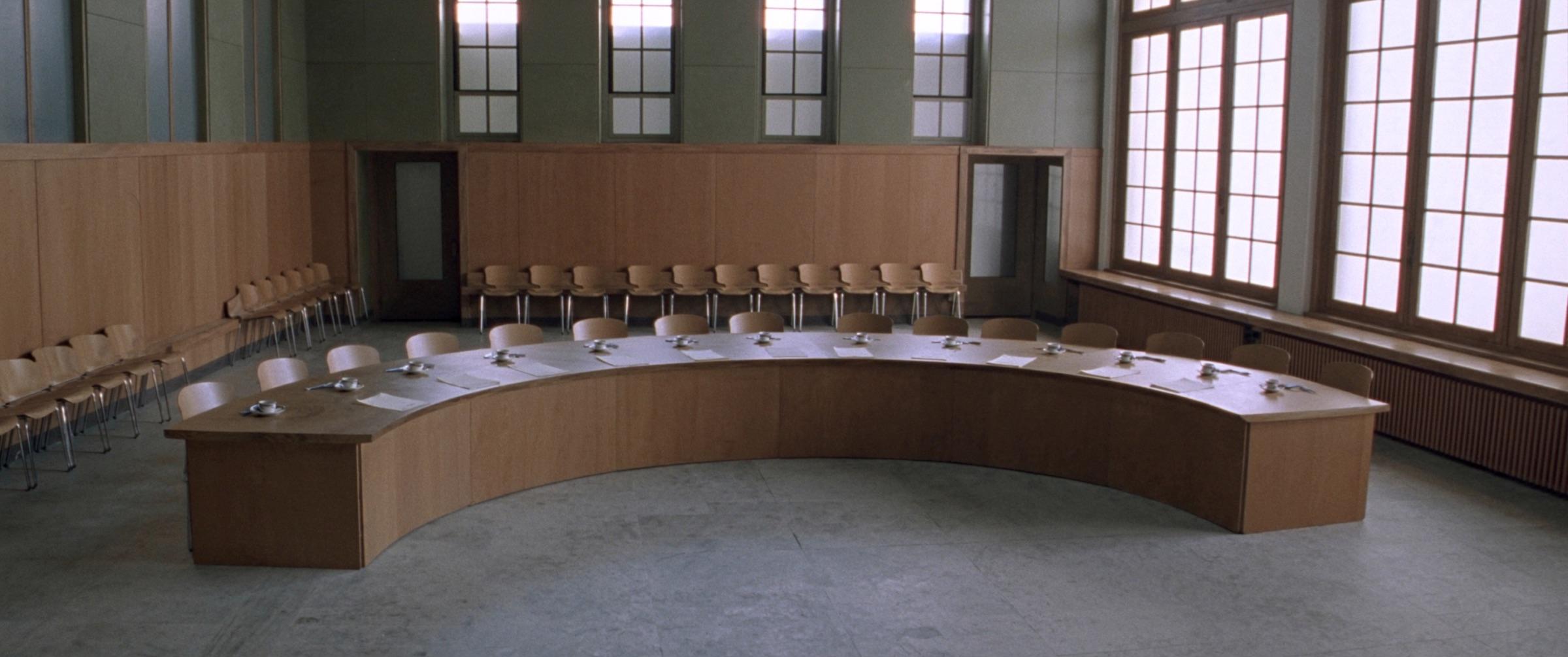 A town hall council chamber: a long table, rows of chairs, tall windows and a stone floor in daylight.